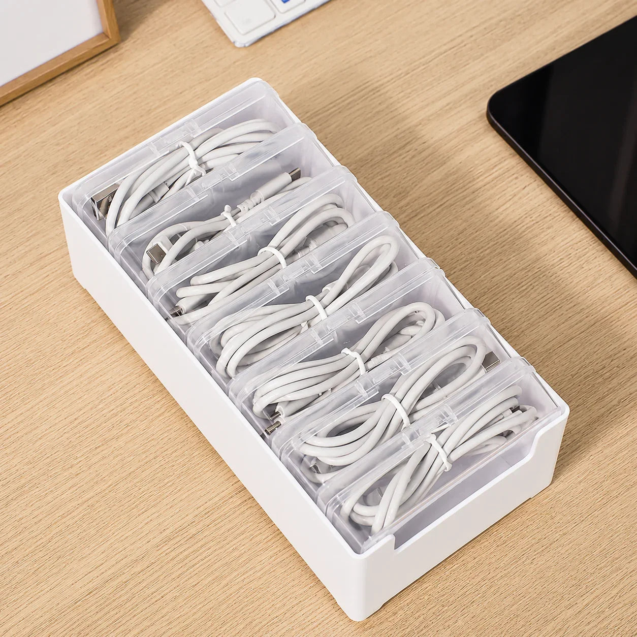 White plastic cable organizer with clear compartments holding neatly coiled white charging cables on wood desk