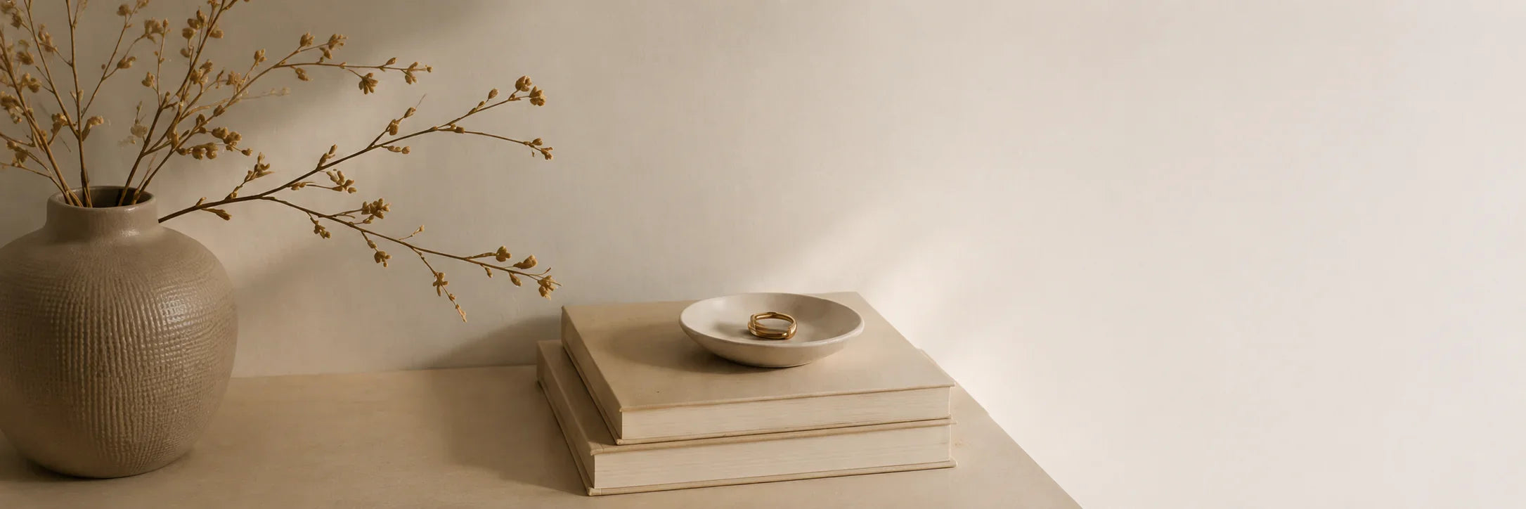 Beige ceramic vase with dried branches beside stacked books and a small dish holding rings