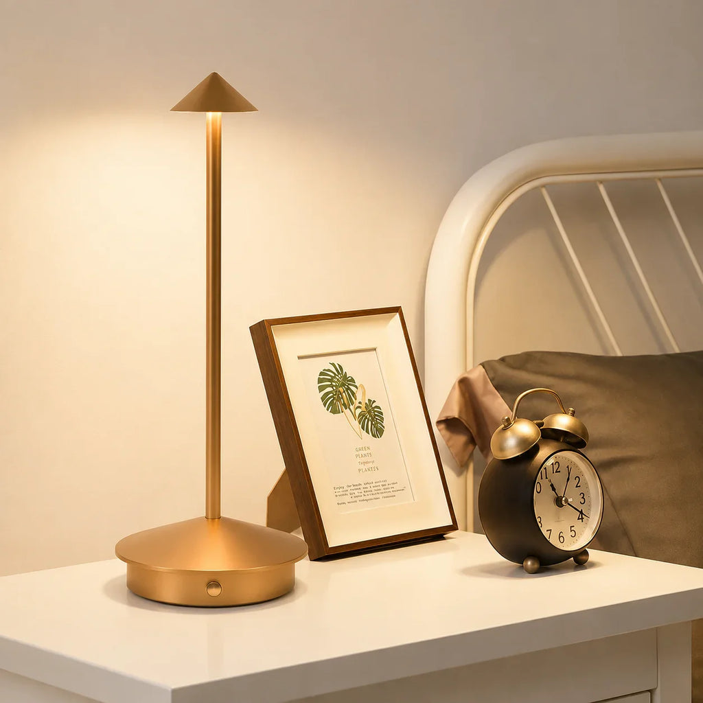 Gold modern table lamp with cone shade on white nightstand beside framed art and alarm clock
