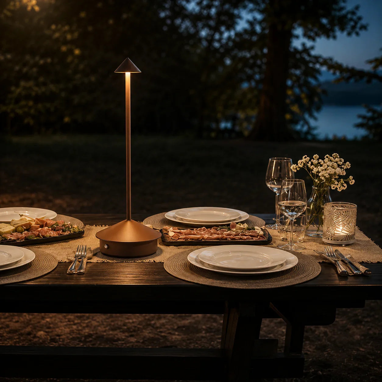 Copper outdoor table lamp illuminating a rustic dinner setup with plates, wine glasses, and charcuterie