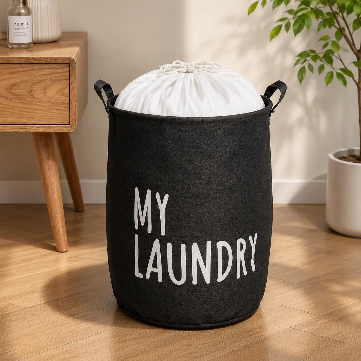 Black fabric laundry hamper with white 'MY LAUNDRY' text, filled with white linens in a home setting.
