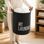 Person holding a black fabric laundry hamper with white 'MY LAUNDRY' text, filled with white linens.