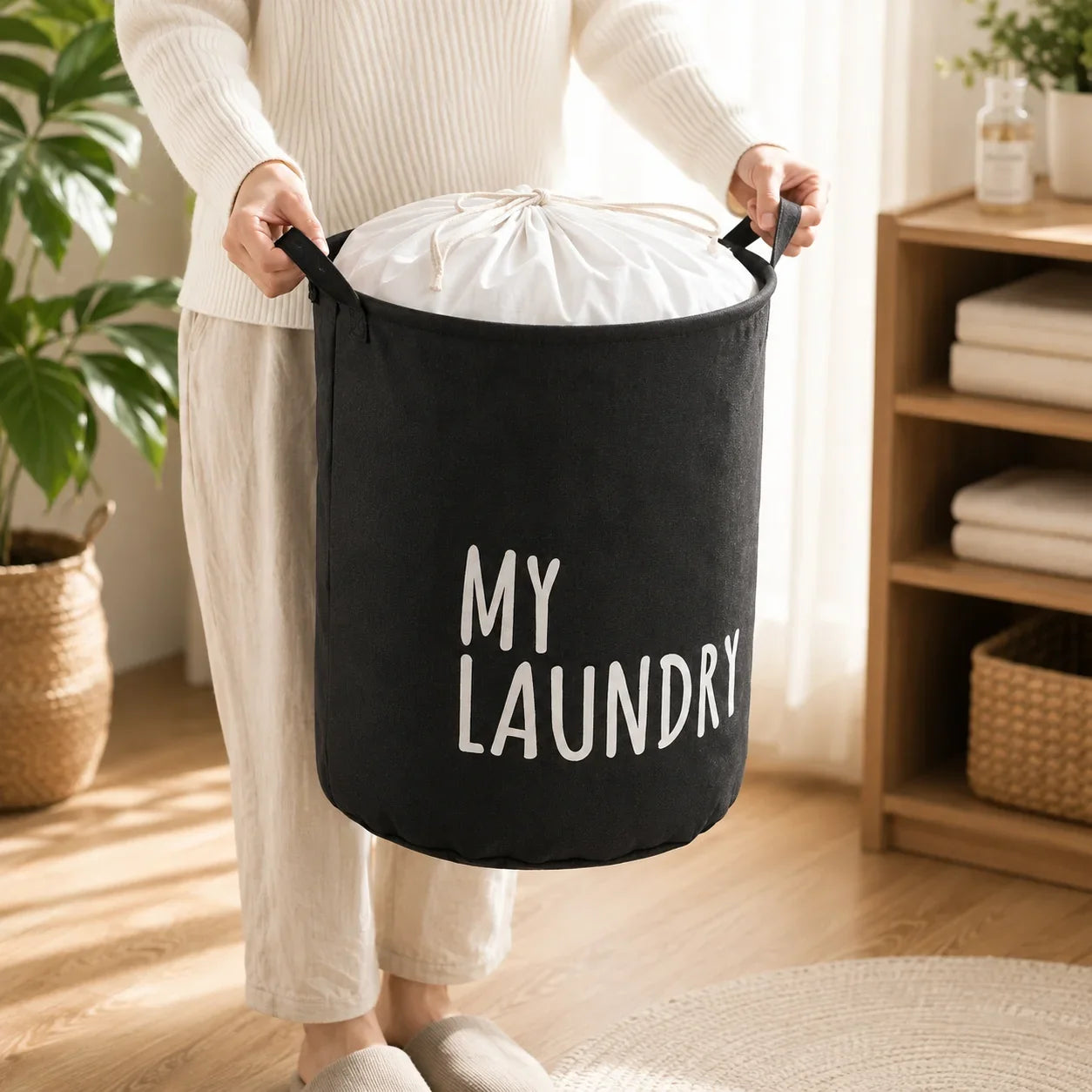 Person holding a black fabric laundry hamper with white 'MY LAUNDRY' text, filled with white linens.