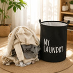 Black fabric laundry hamper with 'MY LAUNDRY' text next to a pile of folded clothes on a jute rug.
