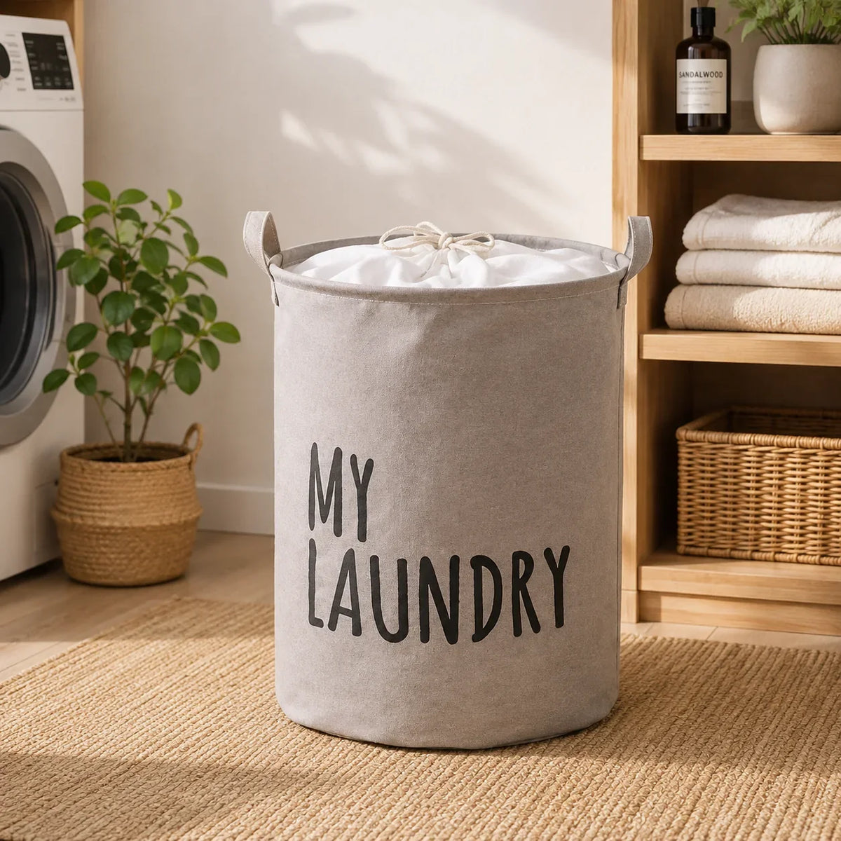 Grey fabric laundry hamper with 'MY LAUNDRY' text and handles, placed on a jute rug in a laundry room.