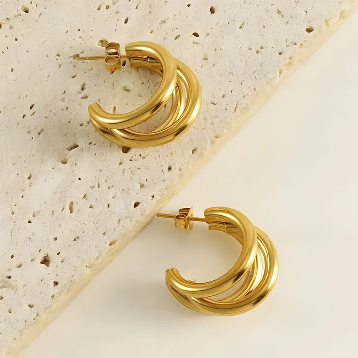 Gold triple hoop earrings, minimalist style, polished finish on textured stone background