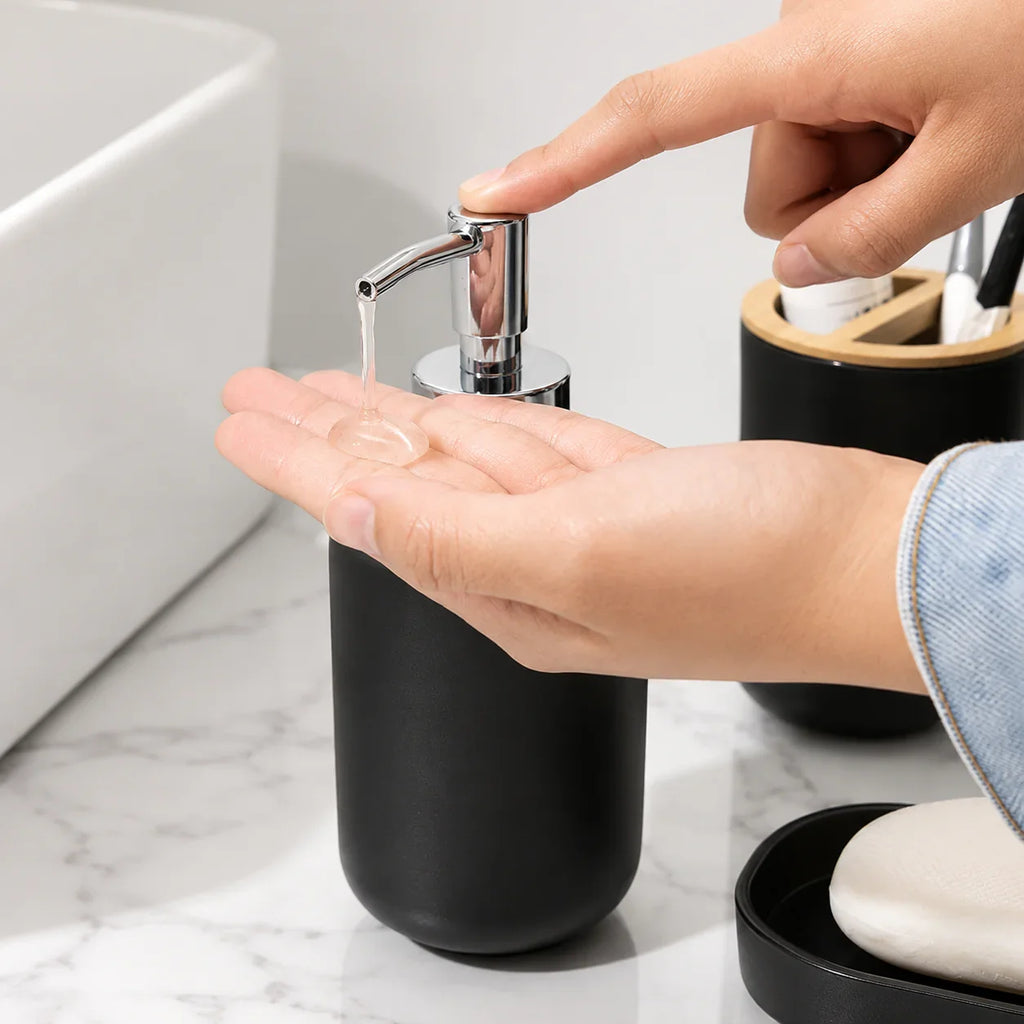 Matte black ceramic soap dispenser with chrome pump dispensing liquid soap into hand on marble bathroom counter