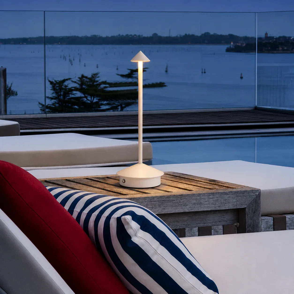 White outdoor table lamp with cone shade on wooden side table beside striped cushion near pool